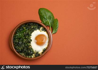 Spinach food with baked eggs in a clay pot on an orange table. Above view with copy space