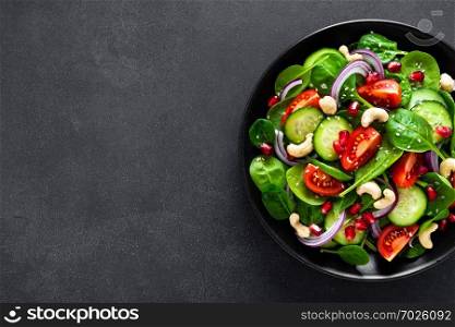 Spinach salad with fresh cucumbers, tomato, onion, pomegranate, sesame seeds and cashew nuts on black background. Healthy vegan food. Top view