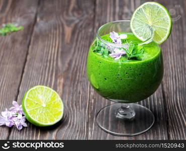 Spinach smoothie with fruits and seeds superfoods, decorated with mint leaves, a slice of lime and wild flowers in a glass cup.Closeup, dark wooden background