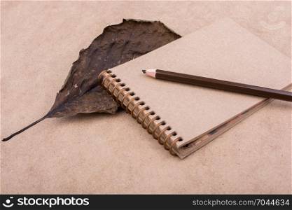 Spiral notebook, color pencil and a dry leaf a brown background