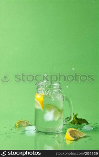 Splash and drops of lemonade from a glass and slices lime, lemon, mint sprig on a green table. Glass jar with homemade sparkling lemonade on green background with copy space.. The cube of ice fell into a glass jar with a splash and drops of lemonade in different directions. Drops of liquid and pieces of lemon, lime on a green table