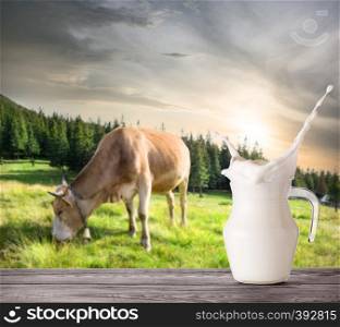 Splash in a jug of milk on a beige cow background. A jug of milk stands on a wooden table against the background of a beige cow on a mountain pasture in the evening. Splash in jug of milk on beige cow background