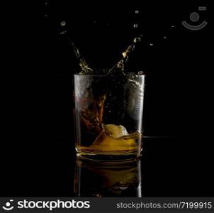 Splash in glass of whiskey and ice block falling on black background