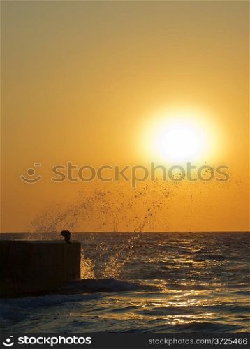 Splashes of sea wave breaking against concrete moorage at sunset.