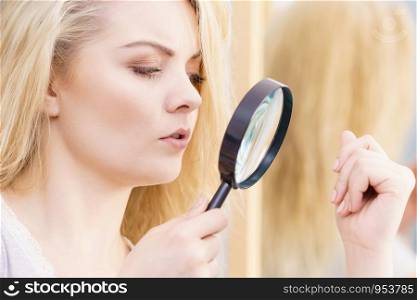 Split ends problem, dry effect, haircare concept. Unhappy blonde woman looking at destroyed damaged hair through magnifying glass. Woman magnifying her split ends hair