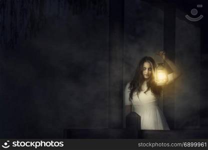Spooky image of a scary woman with dark eyes and appearance of a witch, in a white dress, holding a lit lantern, in a dark night atmosphere.