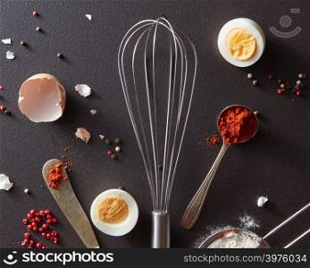 Spoon with red pepper, metal halo, vintage knife, boiled egg and shell on a black concrete background. Concept cooking. Flat lay. Metal whisk, spoon with red pepper knife and halved boiled eggs on a black concrete background. Cooking Concept. Flat lay