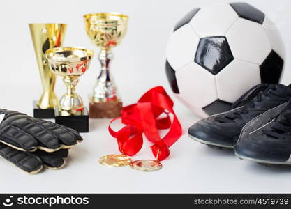 sport, achievement, championship and success concept - close up of soccer ball, football boots and goalkeeper gloves with golden medals and cups