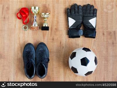 sport, achievement, championship and success concept - close up of soccer ball, football boots and goalkeeper gloves with golden medal and cups on wooden background