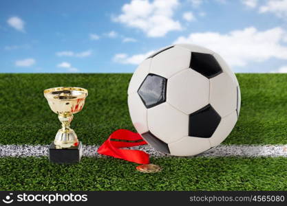 sport, achievement, championship, competition and success concept - close up of football or soccer ball with golden medal and cup over playing field and blue sky background
