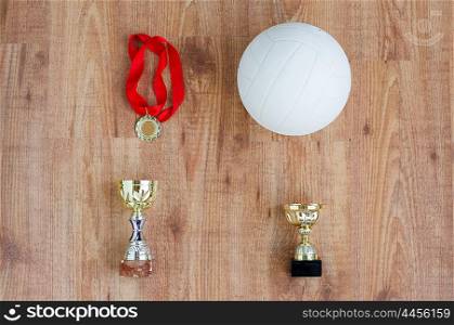 sport, achievement, championship, competition and success concept - close up of volleyball ball with golden medal and cup over wooden background