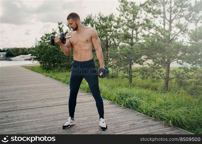 Sport and bodybuilding concept. Muscular bearded European man raises heavy barbells wears sport trousers and sneakers flexes muscles outdoors, does weightlifting exercises, leads active lifestyle