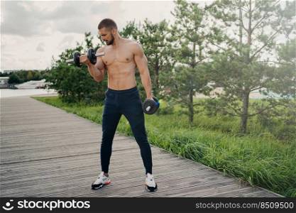 Sport and bodybuilding concept. Muscular bearded European man raises heavy barbells wears sport trousers and sneakers flexes muscles outdoors, does weightlifting exercises, leads active lifestyle