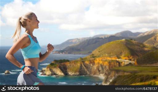sport and healthy lifestyle concept - smiling young woman with fitness tracker running over big sur hills and pacific ocean background in california. woman with fitness tracker running outdoors. woman with fitness tracker running outdoors