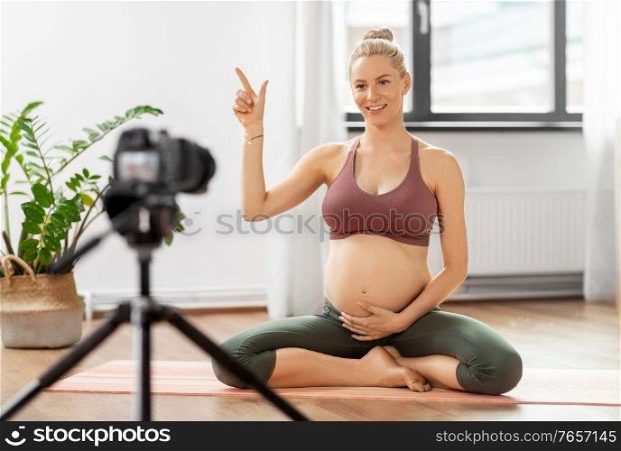 sport and people concept - happy pregnant woman or blogger with camera on tripod recording online yoga class and pointing finger up at home. pregnant woman or yoga blogger with camera at home