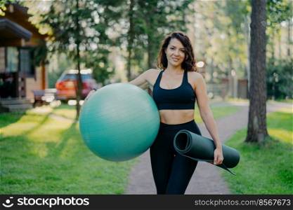 Sport and recration concept. Pleased slim brunette woman wears cropped top and leggings, holds fitball under arm, rolled up karemat, poses outdoor against nature background, stands near trees
