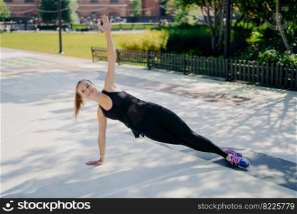 Sport and recreation concept. Athletic slim woman stands in sideways plank raises arm smiles positively dressed in active wear leads healthy lifestyle strengthens core muscles improves stability