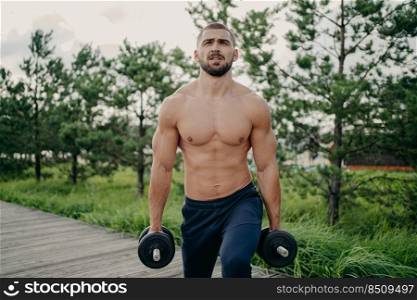 Sport and recreation concept. Muscular unshaven sporty man holds heavy barbells in hands, poses with naked torso, has strong biceps, concentrated into distance, poses outdoor, makes weightlifting