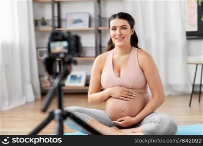 sport, blogging and people concept - happy pregnant woman or blogger with camera on tripod recording online yoga class at home. happy pregnant woman blogger recording yoga video