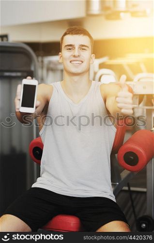 sport, bodybuilding, lifestyle, technology and people concept - smiling young man showing smartphone and thumbs up gesture in gym