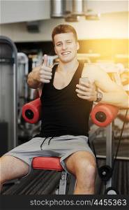 sport, bodybuilding, lifestyle, technology and people concept - smiling young man with smartphone showing thumbs up gesture in gym