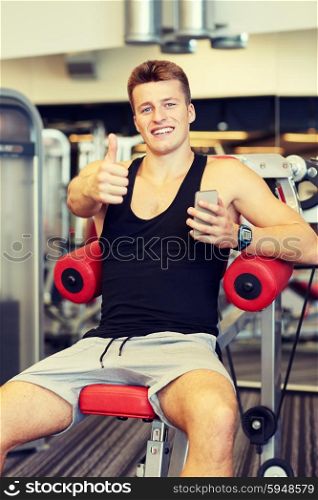 sport, bodybuilding, lifestyle, technology and people concept - smiling young man with smartphone showing thumbs up gesture in gym