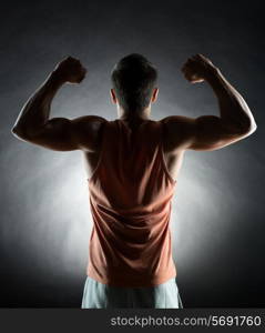 sport, bodybuilding, strength and people concept - young man showing biceps over black background from back