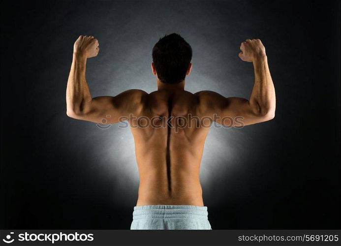 sport, bodybuilding, strength and people concept - young man standing over black background from back and flexing muscles