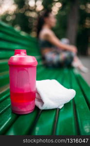 Sport bottle and towel on a bench in summer park, woman in sportswear relax on background. Sport bottle and towel on a bench in summer park
