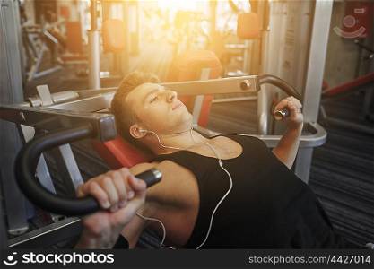 sport, equipment, technology, lifestyle and people concept - young man with earphones flexing chest muscles on gym machine