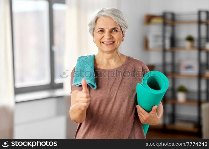 sport, fitness and healthy lifestyle concept - happy smiling senior woman with yoga mat and towel showing thumbs up at home. old woman with yoga mat and towel shows thumbs up
