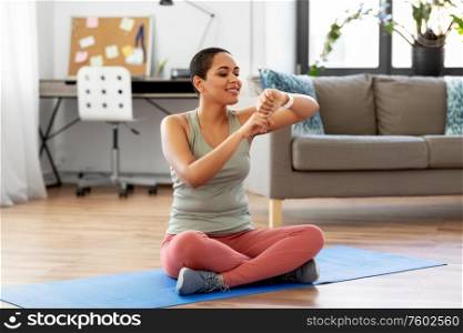 sport, fitness and healthy lifestyle concept - happy smiling young african american woman with smart watch sitting on exercise mat at home. woman with smart watch and exercise mat at home