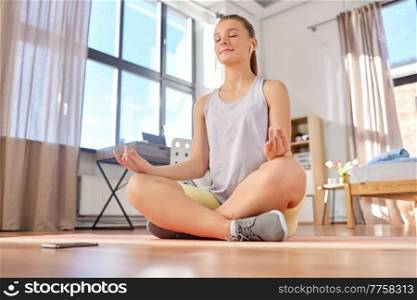 sport, fitness and healthy lifestyle concept - happy teenage girl with smartphone and earphones sitting on yoga mat and meditating at home. girl with phone and earphones meditating at home