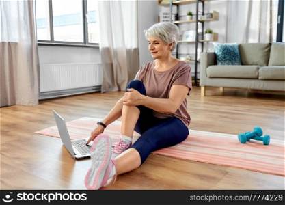 sport, fitness and healthy lifestyle concept - smiling senior woman exercising with laptop computer on mat at home. happy woman with laptop exercising at home