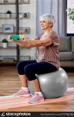 sport, fitness and healthy lifestyle concept - smiling senior woman exercising with resistance band sitting on exercise ball at home. senior woman exercising with elastic band at home