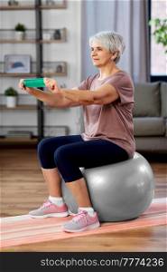 sport, fitness and healthy lifestyle concept - smiling senior woman exercising with resistance band sitting on exercise ball at home. senior woman exercising with elastic band at home