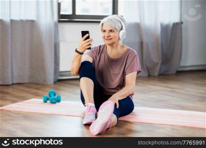 sport, fitness and healthy lifestyle concept - smiling senior woman with smartphone and wireless headphones exercising on mat at home. woman with phone and headphones exercising at home