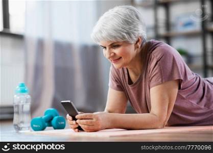 sport, fitness and healthy lifestyle concept - smiling senior woman with smartphone exercising on mat at home. senior woman with smartphone exercising at home