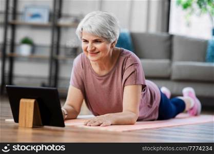 sport, fitness and healthy lifestyle concept - smiling senior woman with tablet pc computer exercising on mat at home. happy woman with tablet pc exercising at home