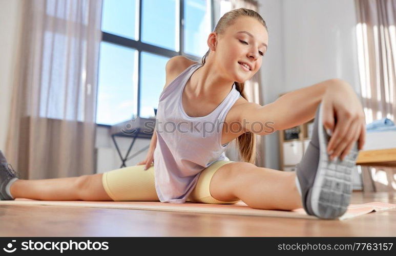 sport, fitness and healthy lifestyle concept - smiling teenage girl exercising on yoga mat and stretching leg at home. happy teenage girl exercising on yoga mat at home