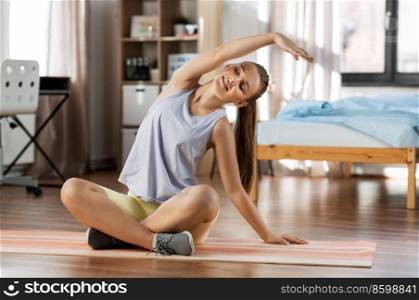 sport, fitness and healthy lifestyle concept - smiling teenage girl exercising on yoga mat and stretching spine at home. happy teenage girl exercising on yoga mat at home