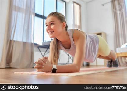 sport, fitness and healthy lifestyle concept - smiling teenage girl with smart watch exercising on yoga mat and doing plank at home. happy teenage girl exercising on yoga mat at home