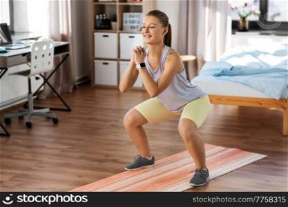 sport, fitness and healthy lifestyle concept - smiling teenage girl with smart watch exercising on yoga mat and doing squats at home. happy teenage girl exercising on yoga mat at home