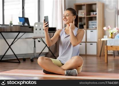 sport, fitness and healthy lifestyle concept - smiling teenage girl with smartphone and earphones sitting on yoga mat at home. girl with phone and earphones exercising at home