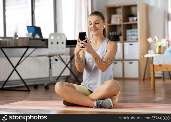 sport, fitness and healthy lifestyle concept - smiling teenage girl with smartphone and earphones sitting on yoga mat at home. girl with phone and earphones exercising at home