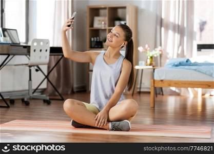 sport, fitness and healthy lifestyle concept - smiling teenage girl with smartphone and earphones sitting on yoga mat and taking selfie at home. girl with phone and earphones takes selfie at home
