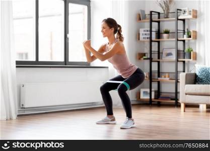 sport, fitness and healthy lifestyle concept - smiling young woman with resistance band exercising and doing squats at home. woman exercising with resistance band at home