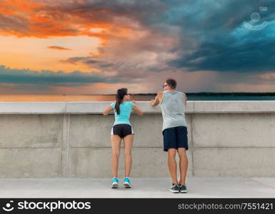 sport, fitness and lifestyle concept - happy couple exercising at concrete wall over sea and sunset sky on background. happy couple exercising outdoors at concrete wall
