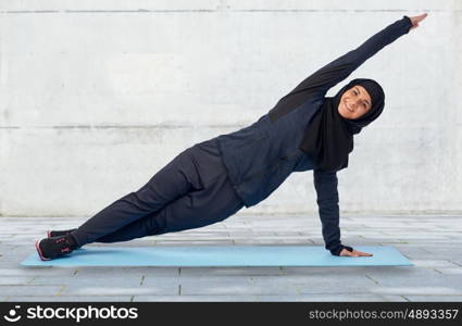 sport, fitness and people concept - happy smiling muslim woman in hijab doing plank exercise on mat over gray concrete wall background
