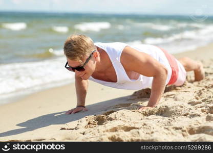 sport, fitness and people concept - happy young man in sunglasses exercising and doing push-ups on summer beach. young man doing push-ups on summer beach